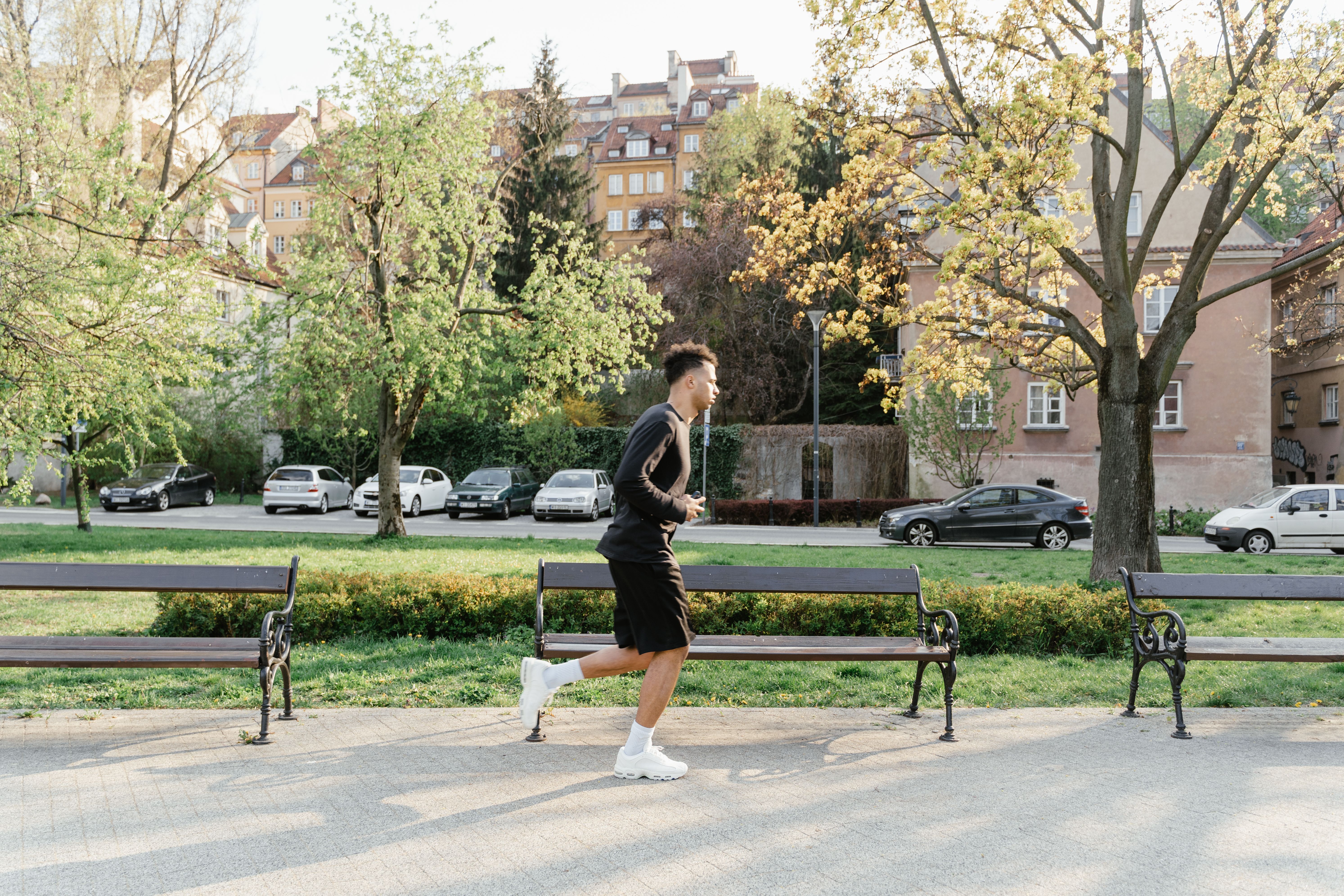 A man in athletic wear jogging through a sunlit park with trees and benches in the background.